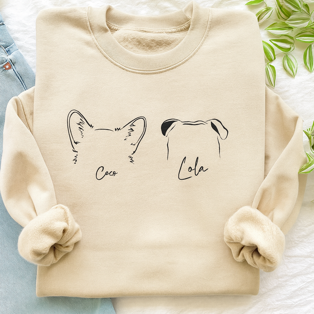 Personalized sand-colored dog sweatshirt featuring custom ear outlines and pet names “Coco” and “Lola,” cozy flat lay with jeans and greenery.