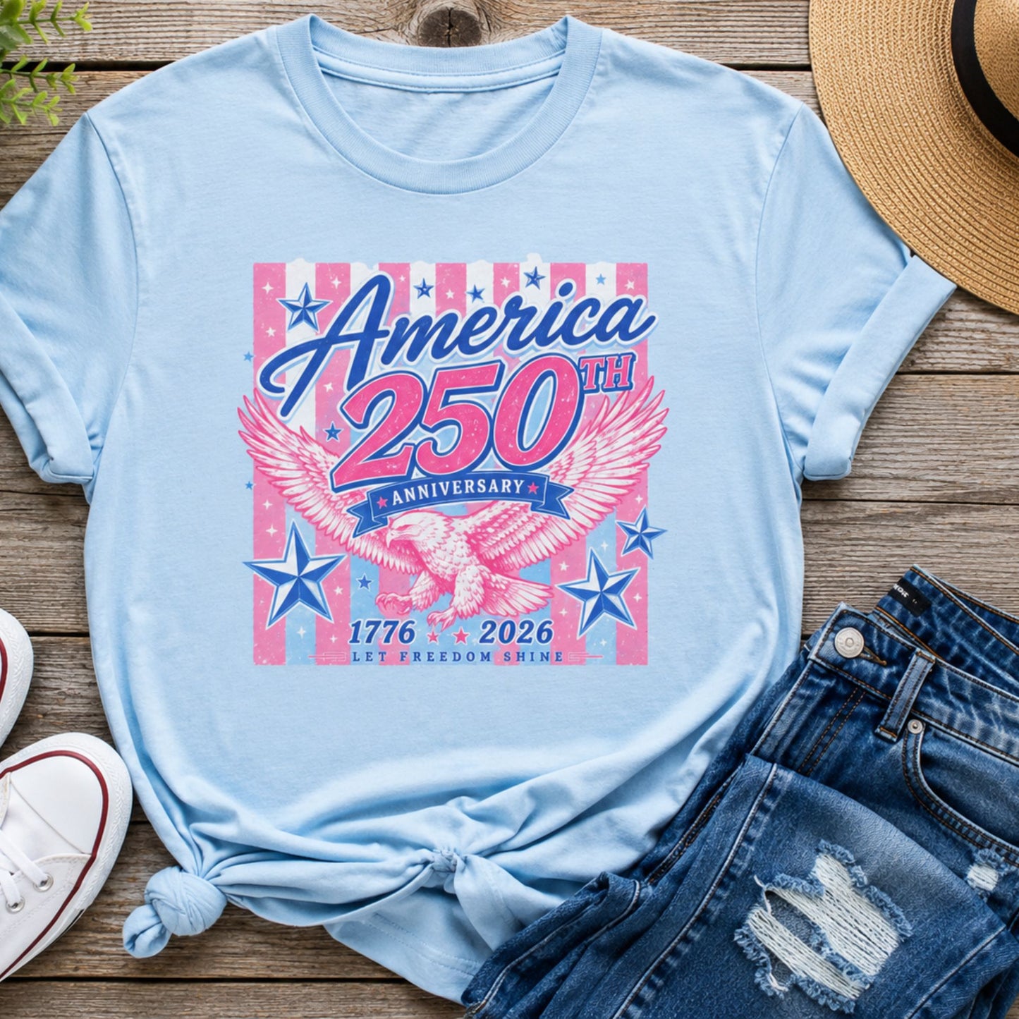 America 250th Anniversary graphic t-shirt in light blue featuring a pink and blue patriotic eagle design with 1776-2026 lettering displayed in a flat lay with denim shorts and white sneakers.