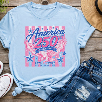 America 250th Anniversary graphic t-shirt in light blue featuring a pink and blue patriotic eagle design with 1776-2026 lettering displayed in a flat lay with denim shorts and white sneakers.