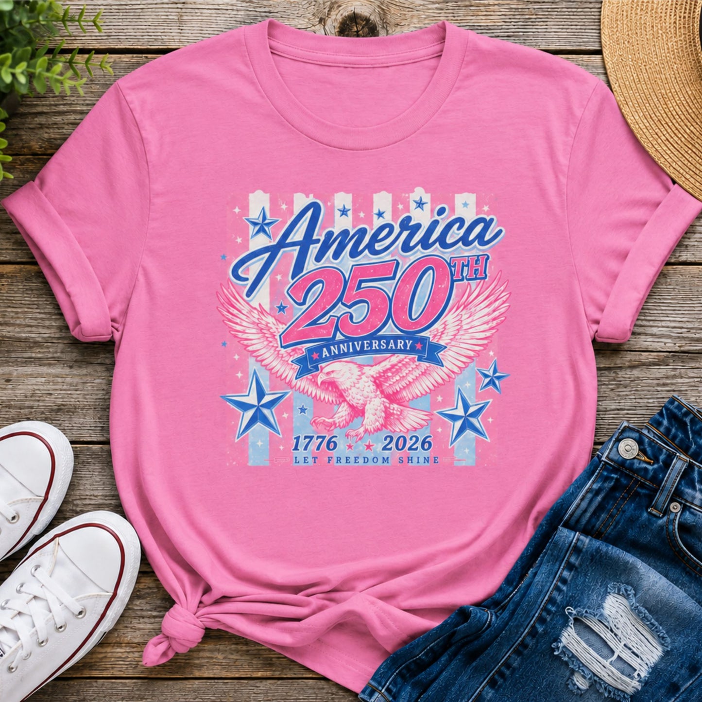 Pink America 250th Anniversary shirt with patriotic eagle graphic, stars, and vintage-inspired 1776-2026 artwork displayed in a summer flat lay with denim shorts.