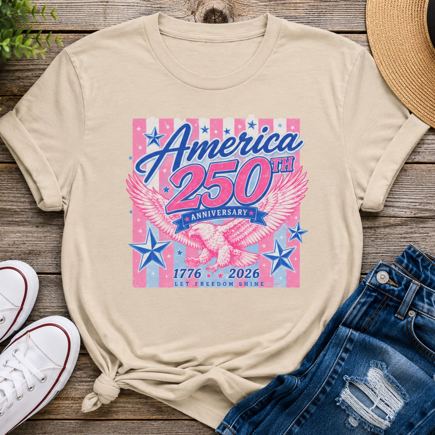 America 250th Anniversary eagle graphic t-shirt in sand color featuring pink and blue patriotic artwork styled with denim shorts and casual summer accessories.