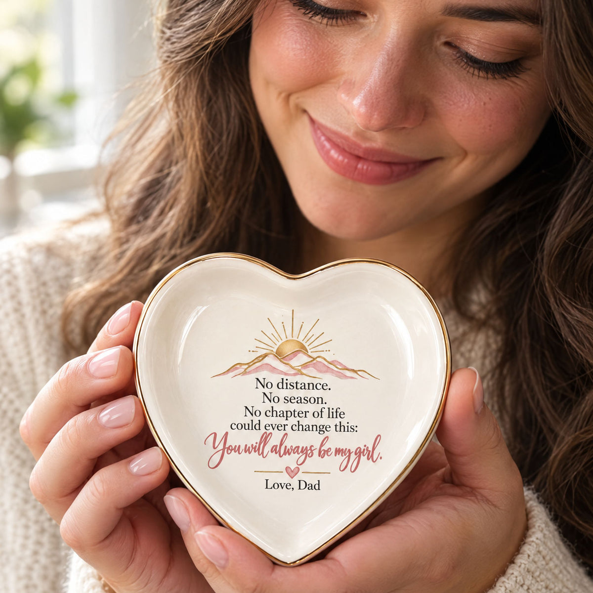 Adult daughter smiling while holding a heart-shaped jewelry dish featuring a loving message signed Love, Dad.
 A woman holds a small heart-shaped ceramic keepsake dish with a gold-trimmed edge and an engraved quote for Dad. The dish is cream-colored with gold detailing and pink script text.