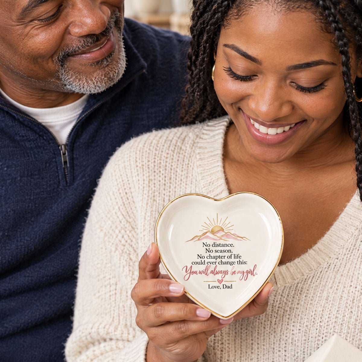 A heart-shaped ceramic keepsake dish with a gold rim and printed inspirational quote is held by a woman while a man in a navy sweater looks on. The dish reads, 'No distance. Smiling father beside his adult daughter as she holds a heart-shaped sentimental jewelry dish gifted from Dad.